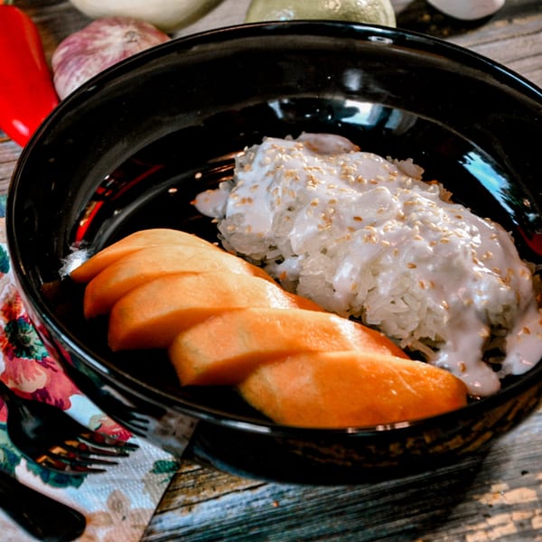 Mango sticky rice with coconut milk and sesame seeds in a black bowl on a wooden table.