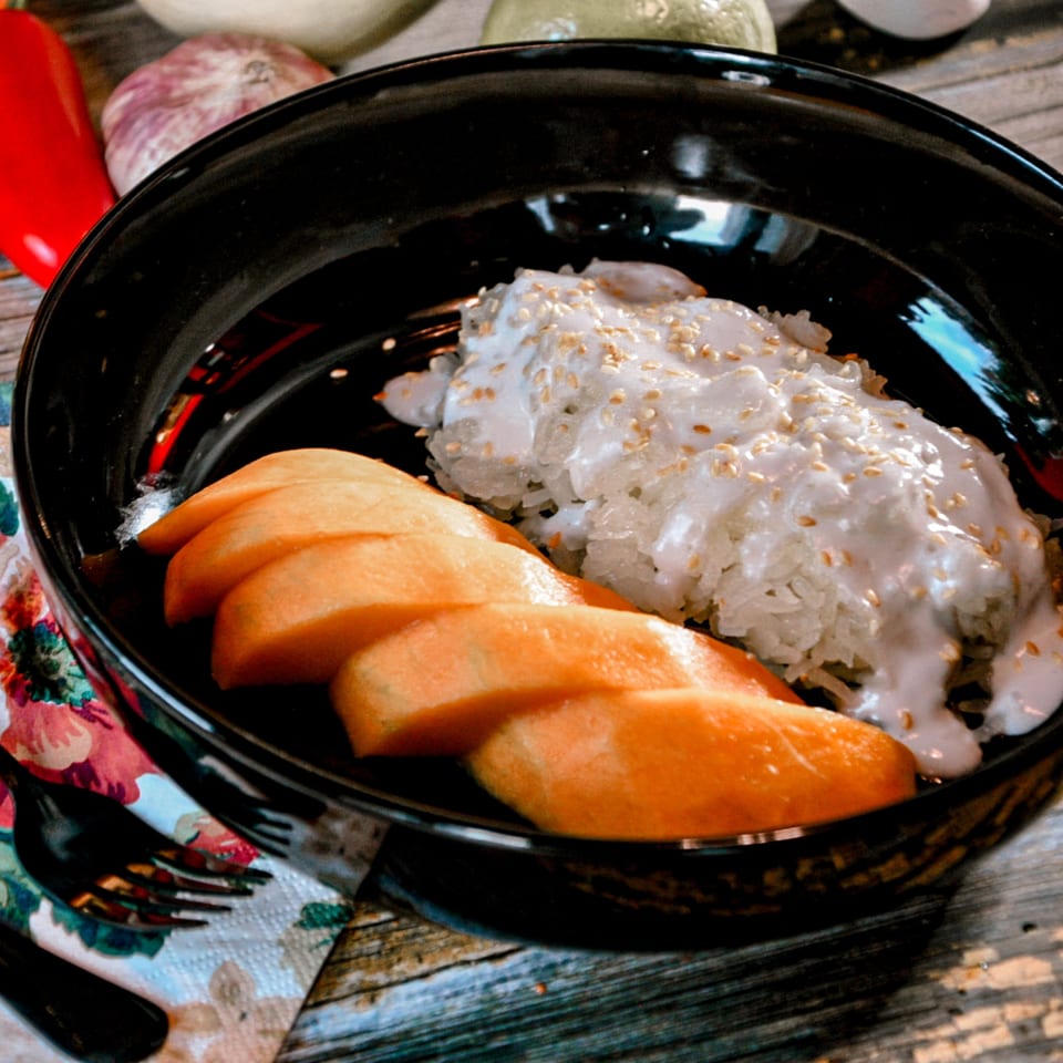 Mango sticky rice with coconut milk and sesame seeds in a black bowl on a wooden table.