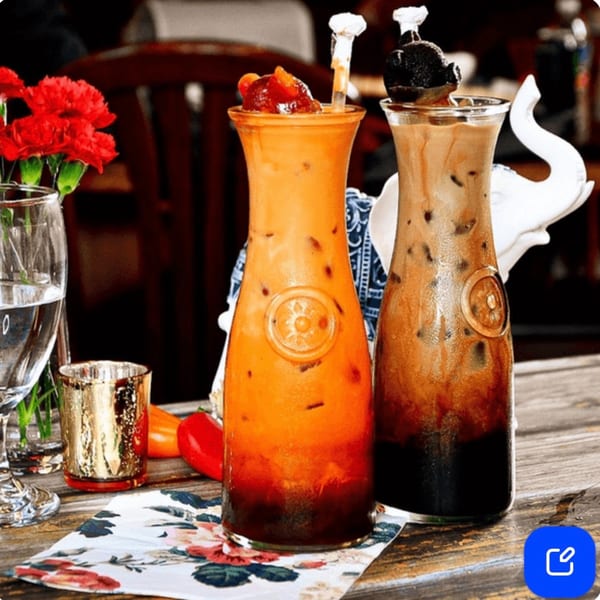 Two tall glasses of iced Thai tea and iced coffee on a wooden table with red flowers.