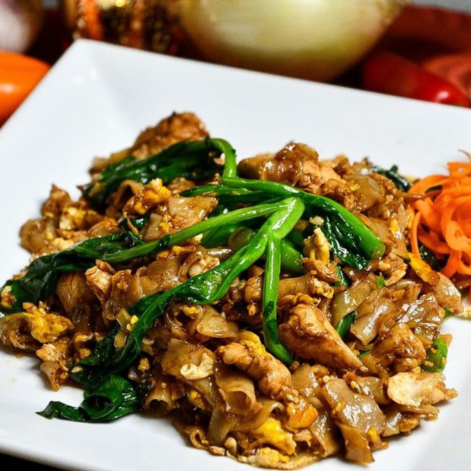 Pad See Ew with wide rice noodles, chicken, Chinese broccoli, and scrambled egg on a white plate.