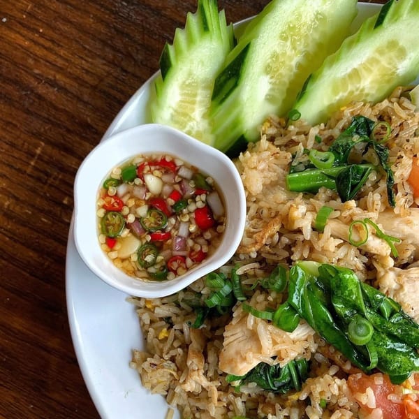 Chicken fried rice with green vegetables, sliced cucumbers, and a chili garlic sauce on a white plate.